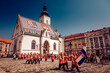 © Travel 'n' Lifestyle - Zagreb, Croatia - 31 August 2019: View of St. Mark's Church with its vibrant tiled roof, as guards in orange uniforms parade with Croatian flag, under a bright blue sky.
