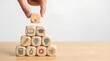 © CuteBee - A hand carefully places a wooden block with a bright lightbulb on top of a pyramid of blocks featuring various education and learning symbols, knowledge, innovation, and academic development.