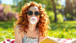 © Athiphat - Curly red haired woman wearing sunglasses blowing bubble gum while sitting on grass in sunny spring park, holding book, relaxed and playful outdoor mood