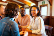 © pressmaster - Multiethnic group of young adults sitting at table in cafe, smiling and talking, biracial woman looking into camera while holding drink, friends enjoying social interaction together