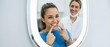 © Fernando - Happy woman patient checking white teeth in mirror at dental clinic, pointing to smile with satisfaction while female dentist watches Concept of oral hygiene, whitening care and stomatology health