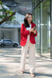 © MINAE - Asian businesswoman in a red blazer and beige pants smiling, walking and holding a mobile phone in her hands on a sunny urban street, managing business communication and digital tasks