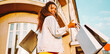 © maxbelchenko - Portrait of a beautiful woman with shopping bags on a city street in the sun's rays. A young woman with a phone enjoys the sunset after shopping. Concepts of consumerism, shopping, and discounts.