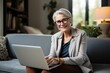 © juliars - Mature woman happily working from home on a laptop, smiling at the camera