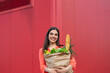 © Graphicroyalty - Portrait of Smiling Woman Holding Large Paper Bag of Fresh Groceries. Young Lady with Healthy Food and Vegetables Standing Against Red Wall. Healthy Eating, Sustainability, and Retail Marketing.