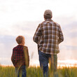 © Acronym - Farmer and his son in front of a sunset agricultural landscape. Man and a boy in a countryside field. Fatherhood, country life, farming and country lifestyle concept.