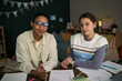 © DragonImages - Portrait of Black young adult woman and Caucasian teenage girl sitting at desk, working together on lesson assignments, holding pens, looking at camera, tutor assisting student