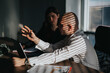 © qunica.com - A business person gestures while discussing at a laptop. Another business employee listens in a sunlit, striped-blind office.