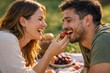 © hobonski - A joyful woman laughs while feeding a fresh red strawberry to her partner during a playful and intimate picnic date in a sun-drenched park.