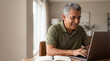 © Николай Срибяник - Senior Hispanic man in green polo shirt smiling while using a laptop at a wooden desk.