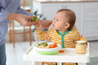 © Pixel-Shot - Mother giving her little baby broccoli in high chair at home