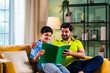 © StockImageFactory - Indian asian son reading a book with father on sofa while preparing for school lessons
