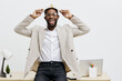© SHOTPRIME STUDIO - Joyful young African American man celebrating birthday or special occasion, holding a party hat on his head and smiling brightly, in casual attire, in an office or home workspace, light background
