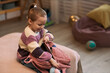 © DragonImages - Caucasian child girl sitting on bed unpacking backpack in cozy room, smiling and looking down while taking out belongings, ball visible in background