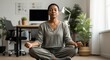 © Nina - Calm African American woman in her 40s practicing mindfulness meditation in lotus pose on the floor of her home office for stress relief and mental health