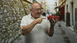 © Krakenimages.com - Senior man holding clear plastic container of strawberries, smiling and giving thumbs up while standing on a narrow stone street beside old building walls; approval summer.