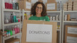 © Krakenimages.com - Woman smiling indoors wearing volunteer uniform holding donation box in charity room filled with supplies and boxes for distribution reflecting community help and support spirit.