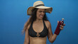 © Krakenimages.com - Woman wearing straw hat and black bikini top grimaces while holding juice bottle in blue studio scene; disgust.