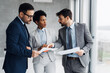 © Novak - Group of three diverse business professionals having a discussion in a modern office, holding and analyzing documents while working on a project together.
