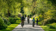 © Broccolini - A happy family of four cycling along a sun-drenched path through a lush green park in spring, surrounded by vibrant flowers and towering trees for a healthy lifestyle.