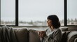 © Feliks - A person in a knitted sweater sits on a couch holding a steaming mug and looks out a large window on a cloudy day A blanket rests beside them