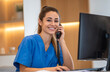 © hestywinarni - Smiling female healthcare receptionist in blue scrubs sitting at desk with computer, talking on landline phone in medical office environment