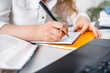 © 5ph - Close up of woman hand writing notes in notebook near open book and laptop, adult student studying online or teacher preparing for distance lesson
