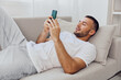 © SHOTPRIME STUDIO - Relaxed man using smartphone on couch, enjoying leisure time with casual outfit and a cheerful mood, surrounded by a bright, modern interior.