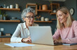 © Pete - Elderly woman learns using laptop with younger woman assisting. Two females interact at table, sharing digital device for online activity. Mature lady types on computer keyboard with help from adult