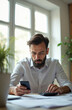 © Viktor - Man with beard reviews papers with phone and calculator. He checks numbers, works on finance in office. Serious worker calculates profit, expense, tax for business.