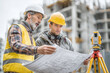 © Thanthara - Construction site planning meeting with two engineers holding blueprint and using surveying equipment for building project outdoors