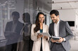 © fizkes - Two smiling teammates, male, female start-up members using tablet stands next to glass black board with written notes, ideas or strategy plan. Teamwork, modern technology, business application usage
