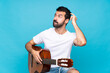 © luismolinero - Young man with guitar over isolated blue background having doubts while scratching head