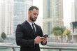 © 24K-Production - Businessman stands in a formal suit, engaged with his smartphone in Dubais urban landscape, representing corporate leadership and success.