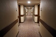 © Gregory - A long, carpeted hotel corridor stretches into the distance with numerous identical wooden doors lining both sides under recessed ceiling lights