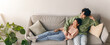 © Prostock-studio - A young couple relaxes on a couch in a living room. They share a moment while the woman leans back comfortably. Plants are visible in the background. Sunlight brightens the space.