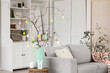 © Pixel-Shot - Tree branches decorated with Easter eggs on stylish table and comfortable grey sofa in living room