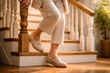 © Elena - Senior Caucasian woman in beige casual outfit standing on wooden staircase at home, wearing orthopedic loafers. Warm natural light, cozy interior, fall prevention and healthy aging lifestyle concept.