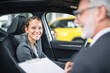 © Kowit - A smiling woman in a car engages with a man in a suit, indicating a friendly conversation, possibly about a vehicle transaction.