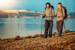 © ivanko80 - Couple enjoys hike along the shore with backpacks and walking sticks near calm water during bright day