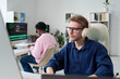 © Mediaphotos - Young man wearing headphones working on computer at desk in office, Black young man sitting at adjacent workstation typing on keyboard, both focused on coding tasks