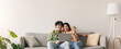© Prostock-studio - A young couple sits together on a couch, using a laptop for online shopping. One person is holding a credit card while smiling. The room is well-lit with plants and a lamp.