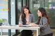© aekachai - Two professional Asian women collaborating at a white desk in a modern office setting. One woman is using a calculator while the other reviews a document intently. They are working together