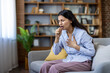 © Liubomir - Unwell young indian woman feeling discomfort in chest, coughing and struggling to breathe, sitting on a sofa at home, experiencing sickness and respiratory illness symptoms