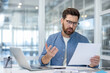 © Liubomir - Man in glasses and beard sitting at a desk, looking at a document with a confused and stressed expression, possibly dealing with difficult financial paperwork in an office environment