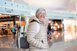 © luciano - Smiling senior caucasian traveler woman with backpack at the airport gate waiting for departure flight, elderly woman holding smartphone looking at camera
