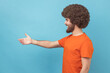 © khosrork - Side view of man with Afro hairstyle wearing orange T-shirt standing with outstretched hand, offering handshake to partner, greeting on job interview. Indoor studio shot isolated on blue background.