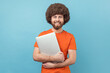 © khosrork - Smiling happy man with Afro hairstyle wearing orange T-shirt standing with notebook in hands, finishing work, looking at camera with positive expression. Indoor studio shot isolated on blue background