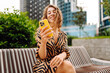 © maxbelchenko - A portrait of a happy woman with a phone stands on a city street overlooking a metropolis. A young tourist texts and enjoys a sunset stroll with her phone. Concepts of technology, urban, and weekend.