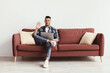 © Prostock-studio - A man sits on a brown couch with cushions, using a laptop and smiling. He waves his hand as if greeting someone. The background is simple and bright.